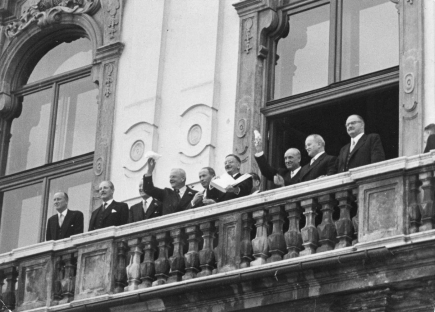 Schwarz-weiß-Foto zur Illustration. Das Foto zeigt Leopold Figl und mehrere Männer in Anzügen auf dem Balkon des Schlosses Belvedere. Leopold Figl hält ein geöffnetes Buch (den Staatsvertrag) in seinen Händen.